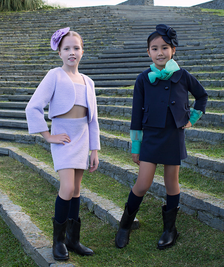 Two stylish girls in coordinated outfits – one in a lilac crop top with matching bolero and skirt, and the other in a navy jacket and skirt ensemble with vibrant green accents, posing on stone steps."